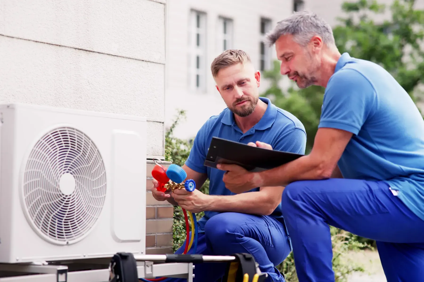 Two HVAC technicians reading a refrigerant gauge set connected to an outdoor air conditioning unit